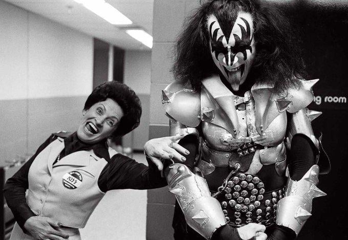 A woman in a KISS-themed uniform poses excitedly next to a smiling person in elaborate, spiked, black-and-white makeup and costume, resembling a member of the rock band KISS, in a backstage hallway.