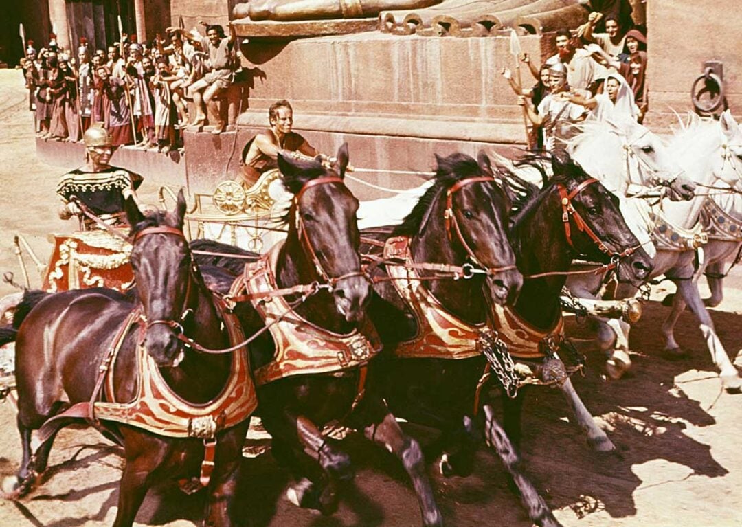 A dramatic chariot race scene with four horses pulling a chariot, a charioteer holding the reins, and a crowd of spectators in ancient-style attire watching and cheering in the background.