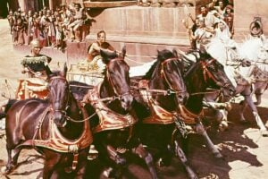 A dramatic chariot race scene with four horses pulling a chariot, a charioteer holding the reins, and a crowd of spectators in ancient-style attire watching and cheering in the background.