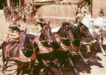 A dramatic chariot race scene with four horses pulling a chariot, a charioteer holding the reins, and a crowd of spectators in ancient-style attire watching and cheering in the background.