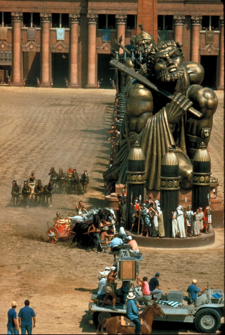 A film set with chariots racing past massive statues of muscular figures holding swords, actors in ancient costumes, large columns in the background, and crew members with cameras and equipment in the foreground.