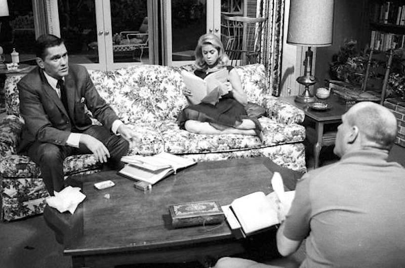 A black-and-white photo shows a man in a suit talking to a woman sitting cross-legged on a floral sofa, reading a book. Another man sits at a coffee table covered with books and papers in a cozy living room.