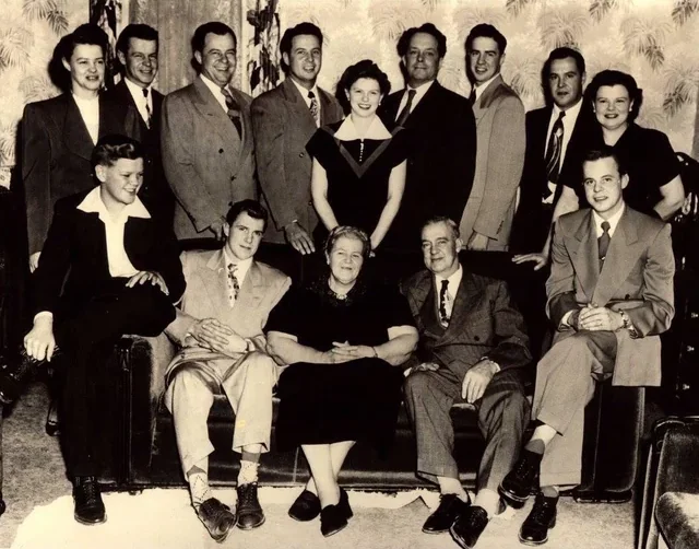 A black and white vintage photo of fourteen people, including men and women, posing indoors. Most are standing behind a sofa, while four are seated in front. Everyone is dressed formally and smiling.