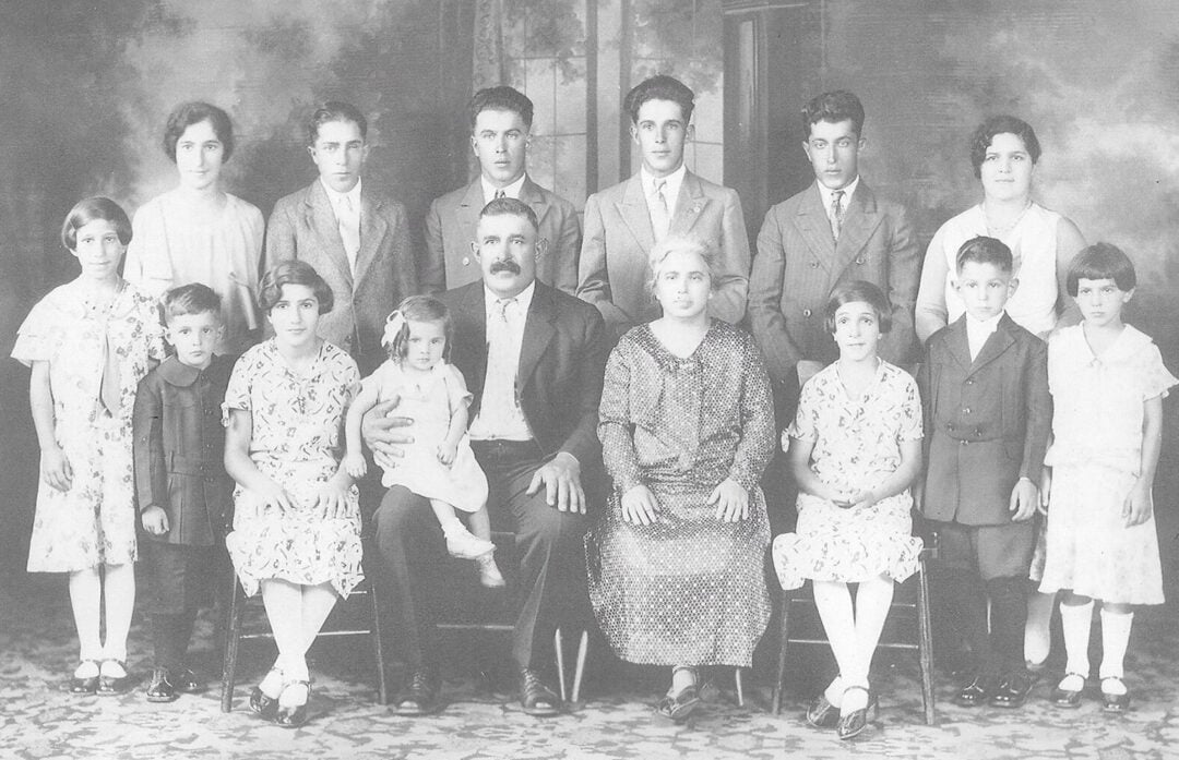 A black-and-white vintage photograph of a large family, with six adults and eight children posing indoors. The adults are seated in the front row, surrounded by children standing and sitting, all dressed formally.