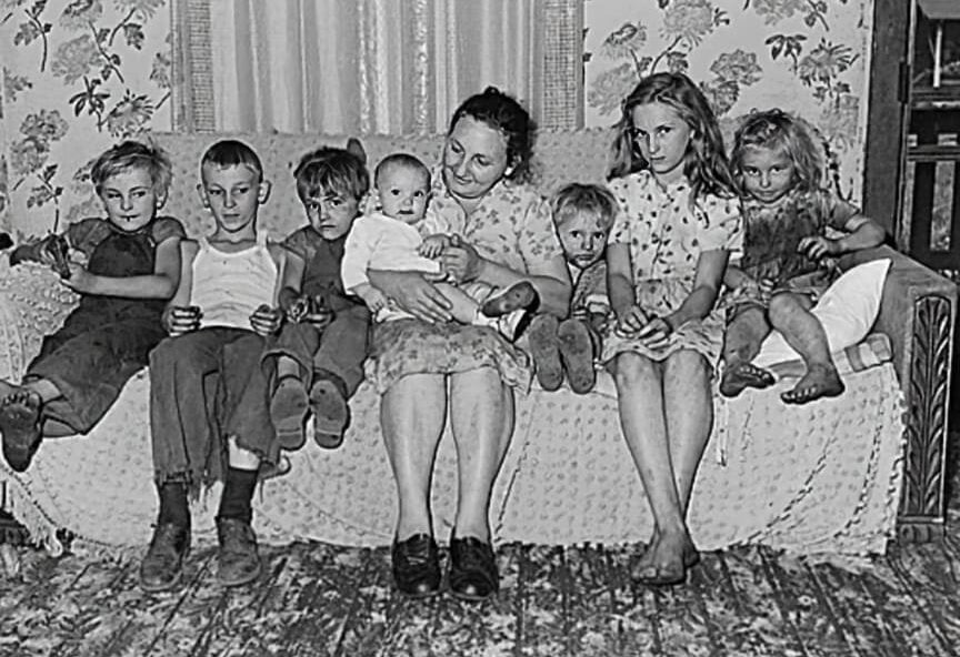 A woman sits on a couch with seven children, all in casual clothes, in a room with floral wallpaper and a wooden floor. The children and woman are closely gathered, some smiling, some looking serious.