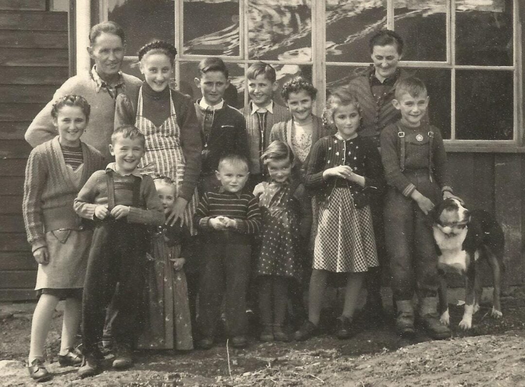 A black and white photo of a group of adults and children standing outside a wooden building, smiling at the camera. A dog is at the front right beside a boy. The adults stand at the back, children in front.