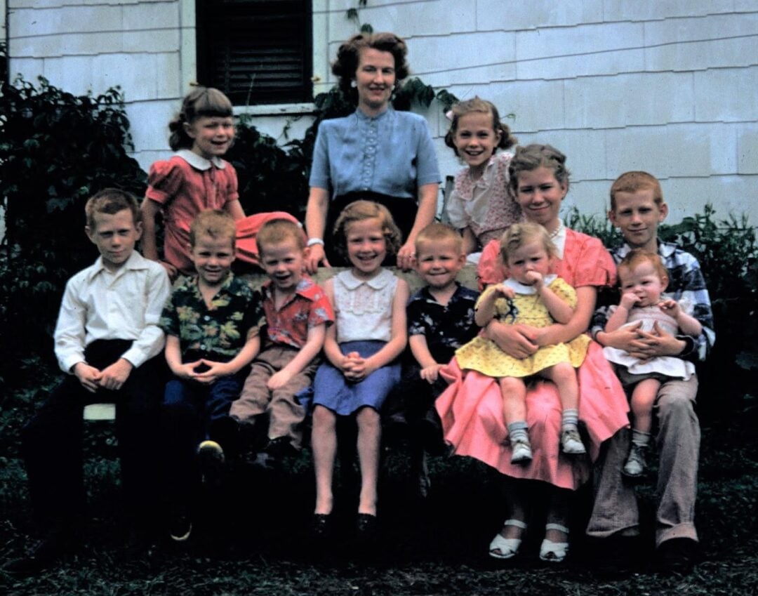 A woman and eleven children of various ages are posed outside in front of a house, dressed in vintage clothing, with greenery around them. The children are seated and standing in rows, smiling at the camera.