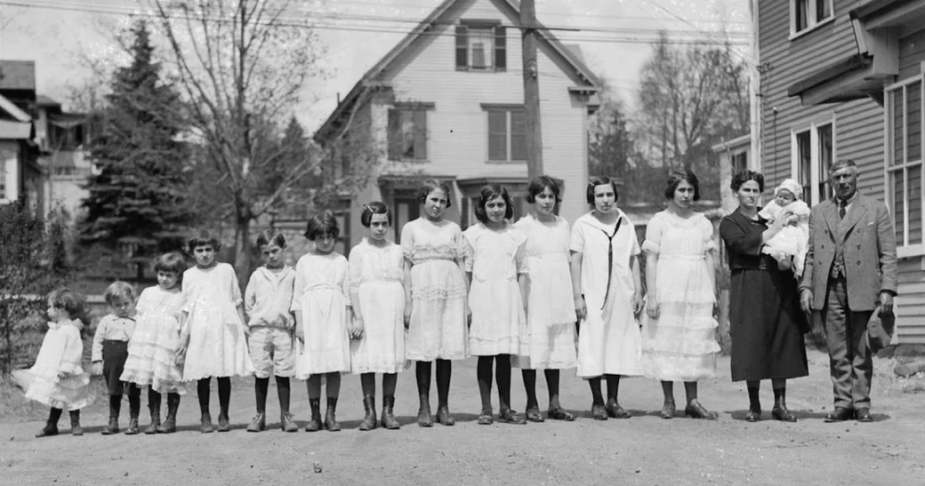 A black and white photo of a large family standing in a row on a dirt road, with fifteen children of various ages and two adults. Houses and trees are visible in the background.