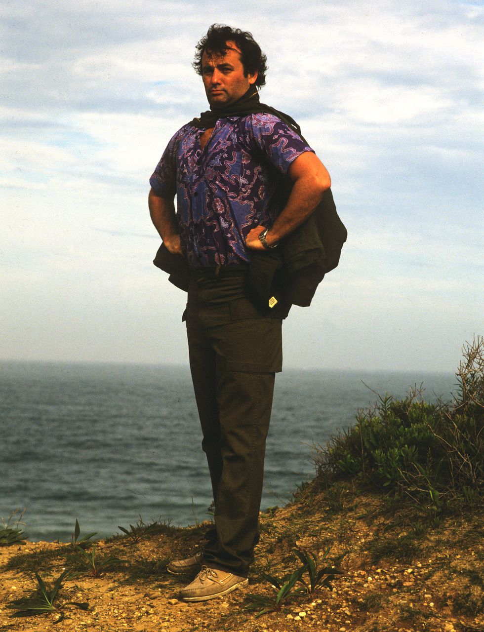 A man stands on a grassy cliff overlooking the ocean, wearing a purple patterned shirt, dark pants, and holding a jacket over his shoulders. The sky is cloudy and the sea stretches out in the background.