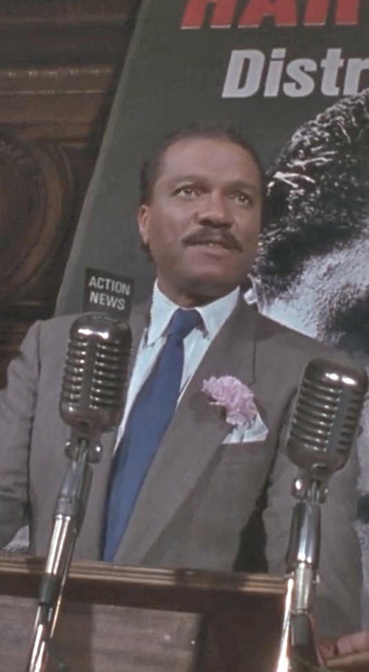 A man in a suit with a blue tie and a pink flower on his lapel stands at a podium with two vintage microphones, speaking, with a partially visible poster and "Action News" sign in the background.