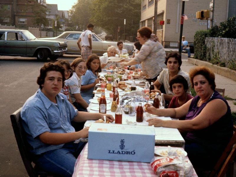 A group of people, including children and adults, sit around a long table outdoors covered with food and drinks, sharing a meal on a residential street. A large Lladró box is in the foreground.