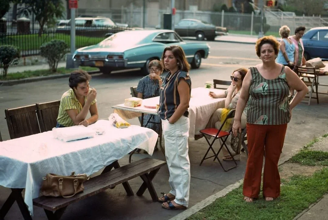 Several women sit and stand around picnic tables covered with white tablecloths on a sidewalk. A vintage blue car is parked nearby, and a street with trees and buildings is in the background.