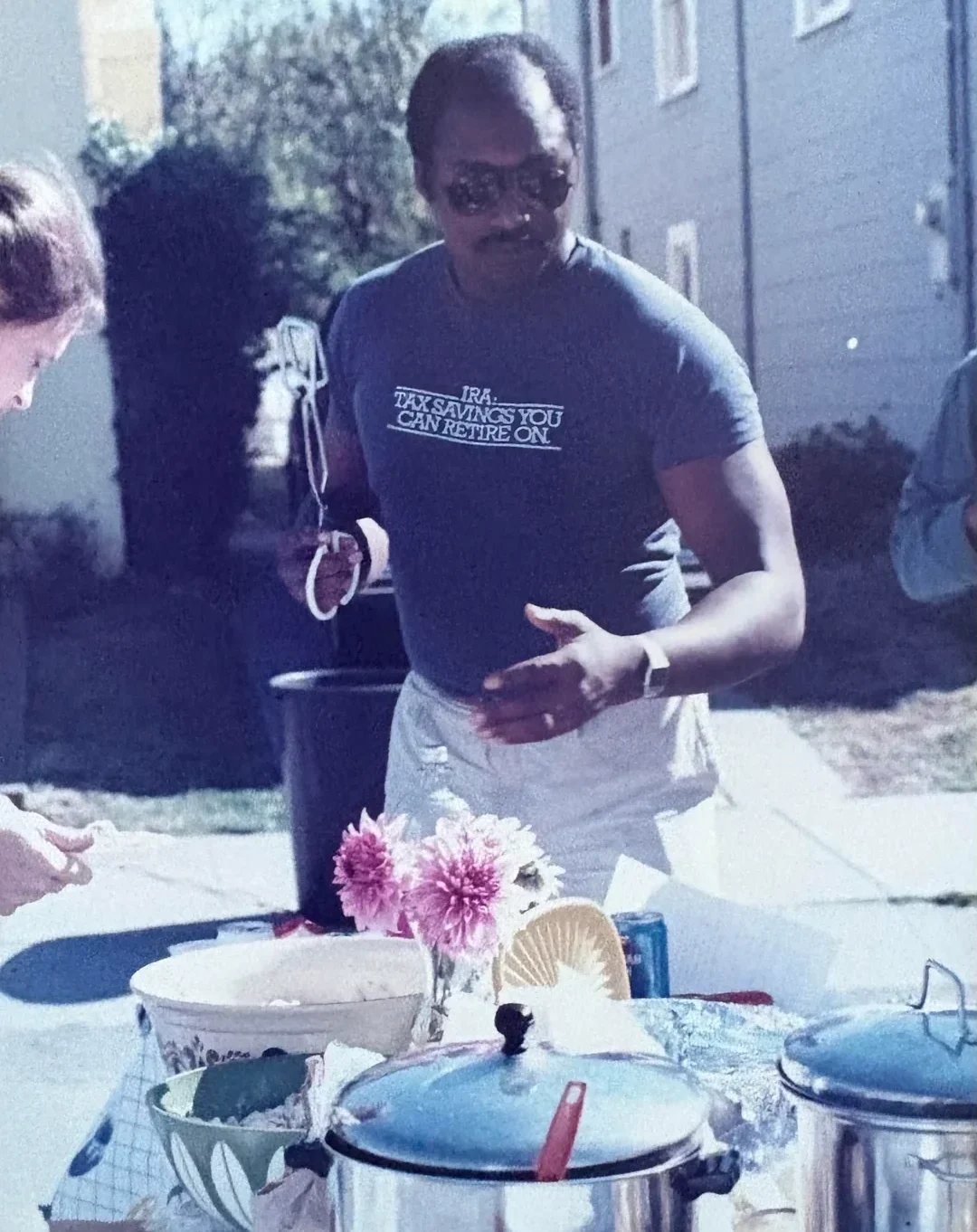 A man wearing sunglasses and a t-shirt that reads “IRA. TAX SAVINGS YOU CAN RETIRE ON” stands outdoors by a table with dishes, flowers, and food, appearing to serve or prepare a meal.