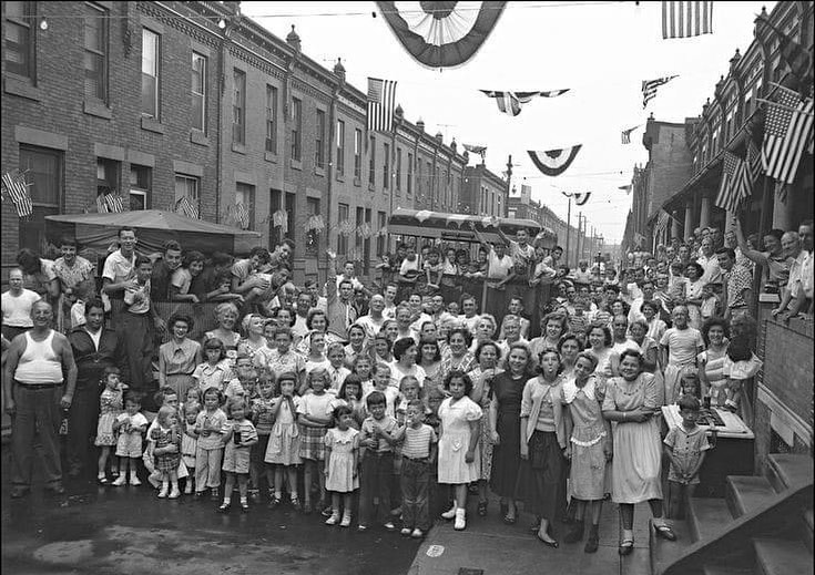 A large crowd of people, including many children, gathers on a city street lined with row houses decorated with flags and banners, likely for a celebration or parade. The mood appears festive and communal.