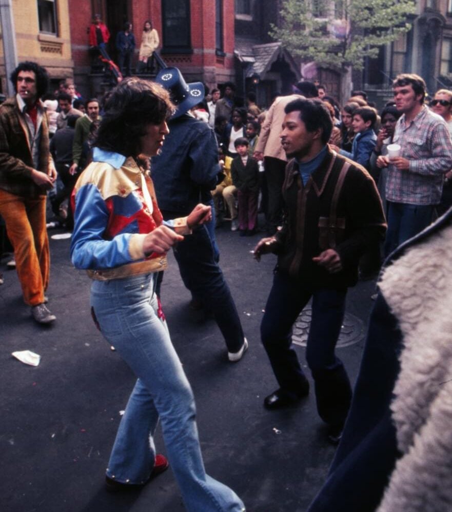 People dance enthusiastically on a city street surrounded by a crowd, with brownstone buildings and trees in the background. The scene appears lively and energetic, evoking a sense of a 1970s block party.