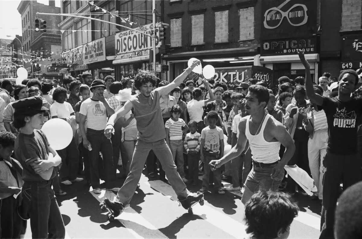 A lively street scene shows a crowd gathered to watch two men breakdancing in the center, while others cheer and hold balloons, during a sunny day in an urban neighborhood.