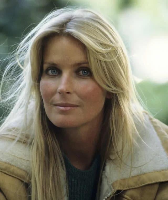 A woman with long blonde hair and blue eyes looks at the camera. She is wearing a tan jacket with a shearling collar over a blue top. The background is blurred and outdoors with greenery.