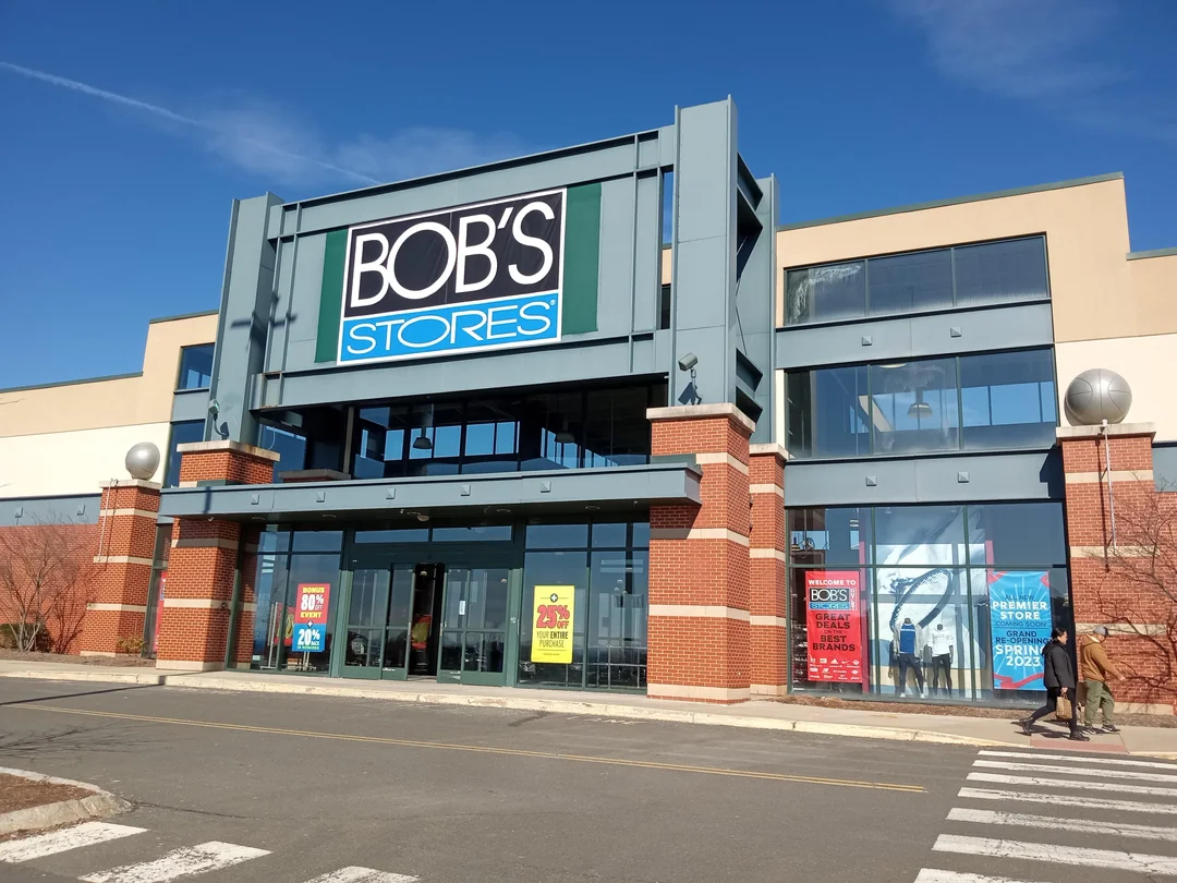 Large Bob’s Stores retail building with tall windows and red brick accents, featuring sale signs in the windows and a clear blue sky above.