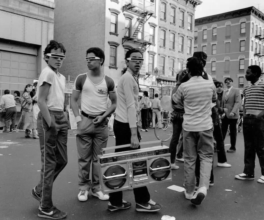 A group of young people stand on a city street in the 1980s, three wearing visor-style sunglasses and tracksuits, one holding a large boombox. Other people and apartment buildings are visible in the background.