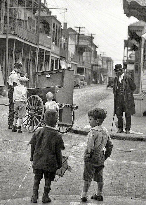 Four children watch a man with a street organ on a cart at a city intersection, while another man in a suit and bowler hat stands nearby; historic buildings and power lines are visible in the background.