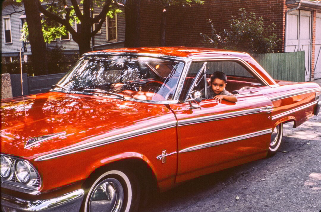 A young boy leans out the driver’s window of a shiny red vintage car parked on a residential street lined with trees and houses.