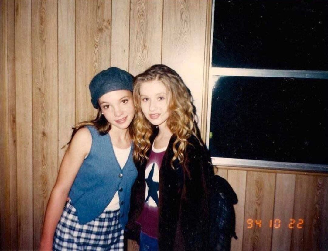 Two teenage girls stand close together indoors, smiling at the camera. One wears a gray cap, blue vest, and plaid skirt; the other has curly hair, a jacket, and a shirt with a star. The wall behind them is wood-paneled.