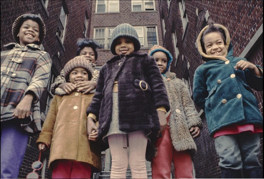 Six children wearing colorful winter coats and hats stand close together, smiling and posing for the camera in front of a tall brick apartment building. The photo is taken from a low angle.