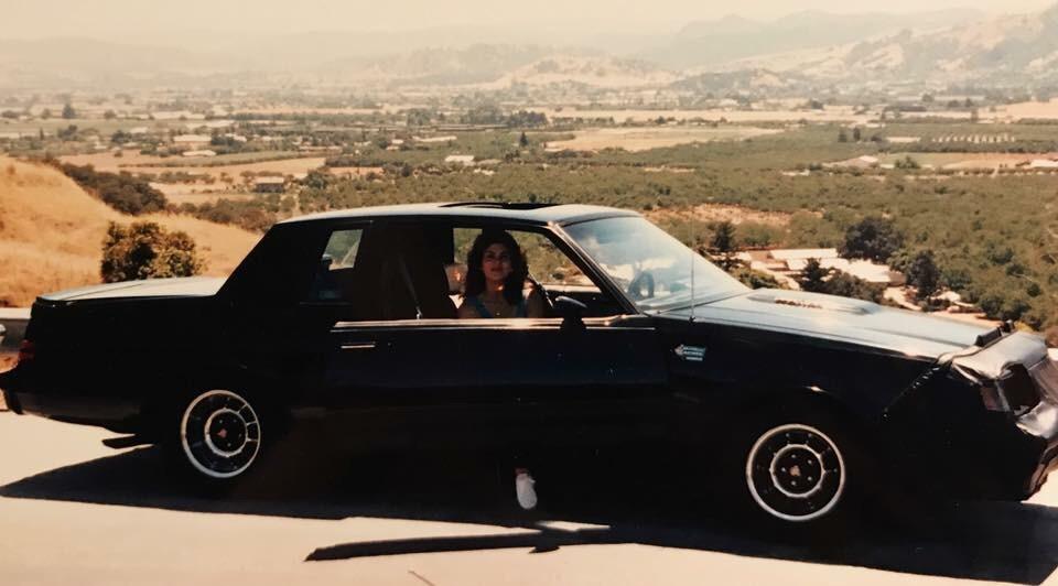 A person sits in the driver's seat of a black classic car with the door open, parked on a hill overlooking a scenic valley with fields, trees, and distant mountains under a clear sky.