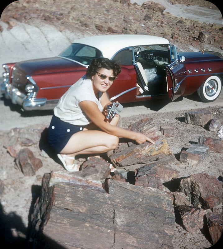 A woman in sunglasses and a white shirt crouches by layered rocks, holding a camera, with a vintage red and white car parked behind her and its door open. The scene is outdoors on rocky terrain.