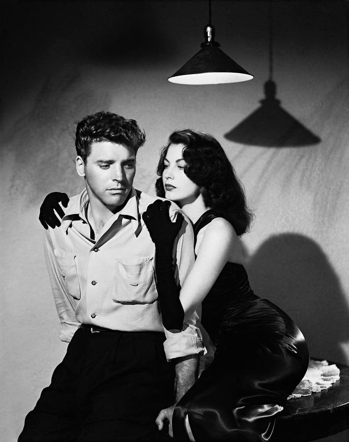 A woman in a black dress and gloves leans seductively on the shoulder of a seated man in a light shirt, beneath a hanging lamp casting dramatic shadows on the wall behind them.