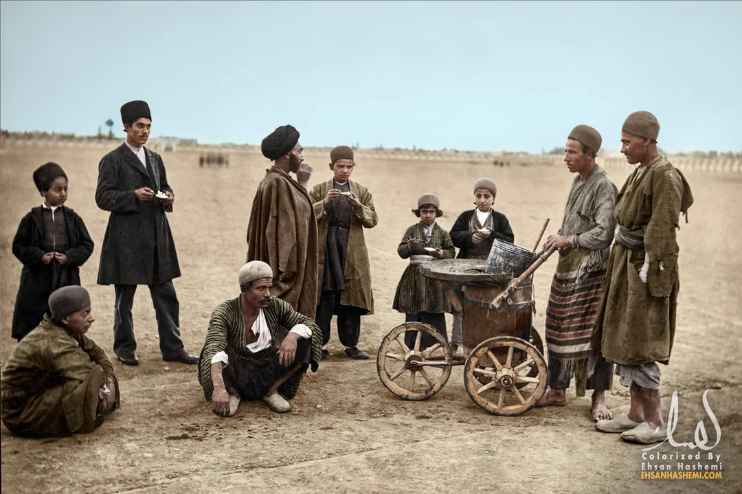 A group of men and boys in traditional clothing gather around a street vendor with a cart in an open, sandy area. Some are standing, others are sitting, and the scene appears historical and outdoors.