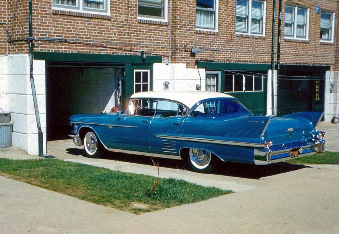 A vintage blue and white 1950s Pontiac car is parked on a driveway in front of a brick building with open garage doors and apartment windows above.