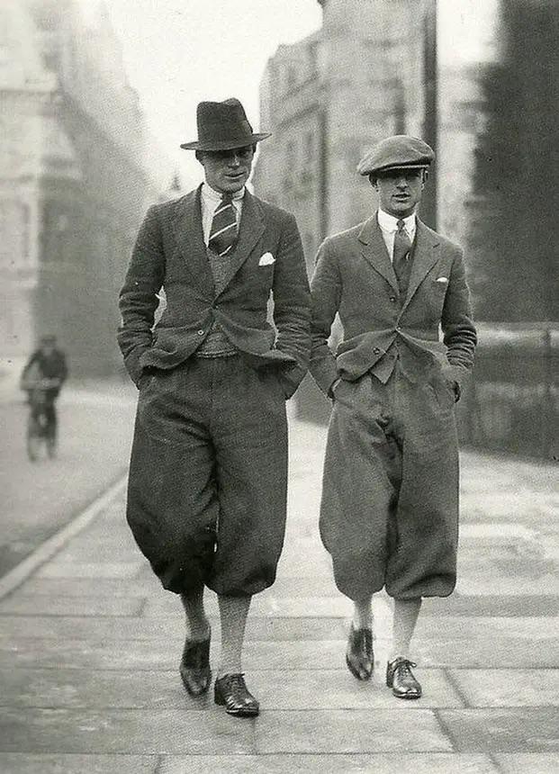 Two men in 1920s attire walk down a city sidewalk. Both wear baggy pants tucked into high socks, jackets, collared shirts, and ties. One wears a fedora, the other a flat cap; a cyclist rides in the background.