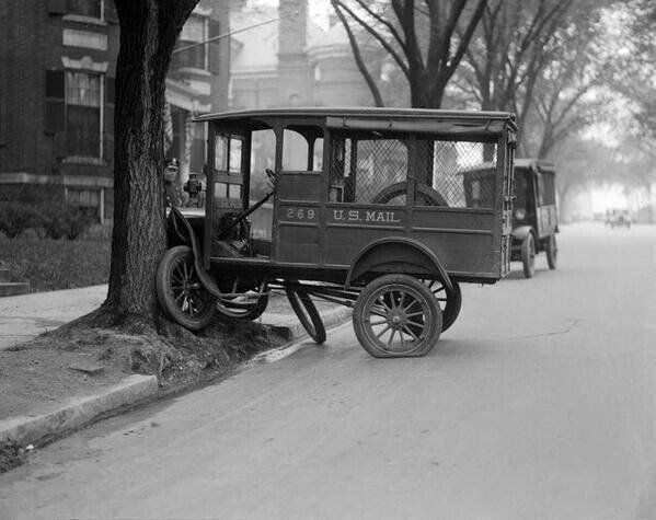 An old-fashioned U.S. Mail delivery truck with a broken front axle leans into a tree on a quiet city street, while another vehicle is parked further down the road.