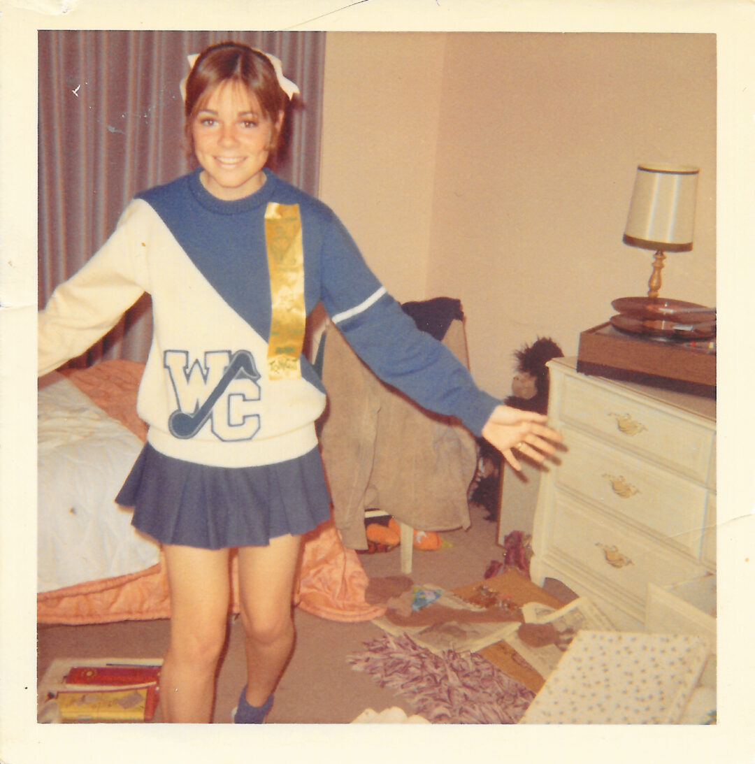 A smiling girl in a blue and white cheerleader outfit with "WC" on the sweater stands in a cluttered bedroom with an unmade bed, dresser, lamp, and scattered items on the floor.