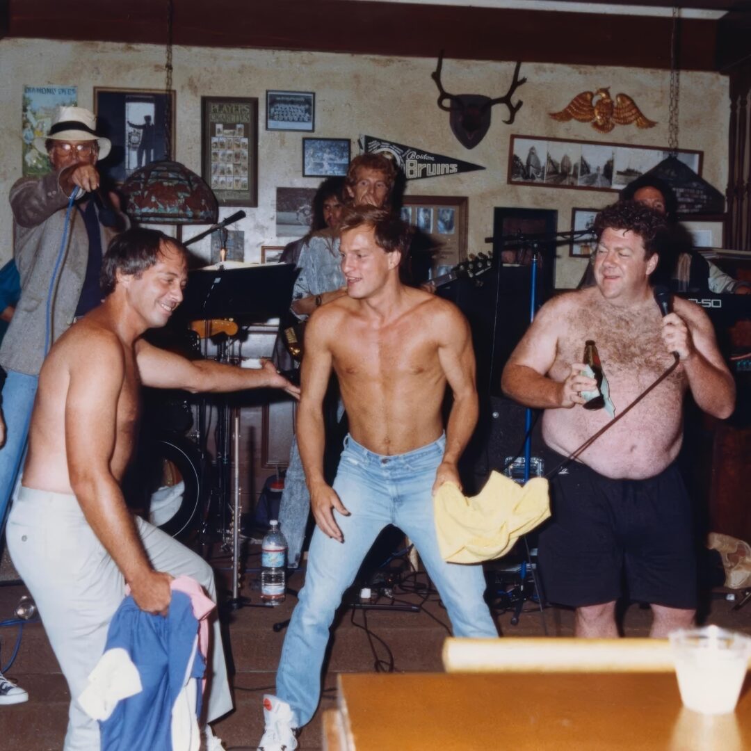 Three men, two shirtless, dance and laugh together in a lively bar, with a live band playing in the background. One man holds a beer bottle. The bar walls are decorated with various memorabilia and pictures.