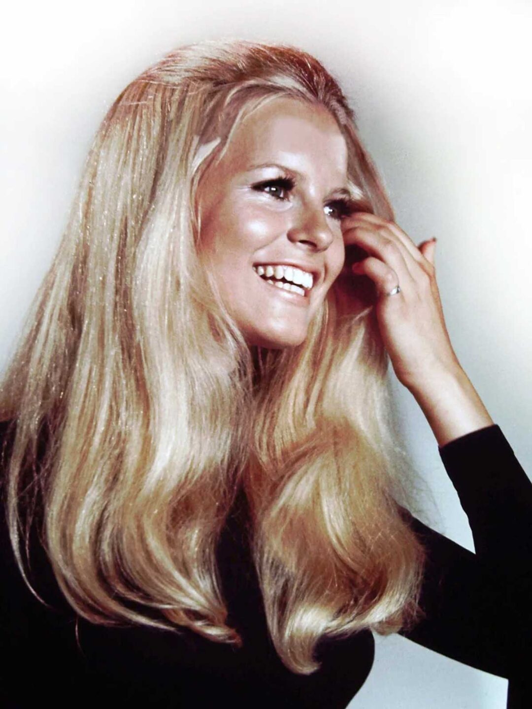 A smiling woman with long, voluminous blonde hair wears a black top and touches her hair with one hand. The background is plain and light-colored.