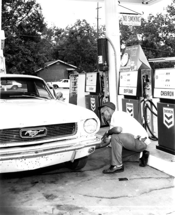 A man in a uniform crouches by the front bumper of a classic Ford Mustang at a Chevron gas station, with vintage gas pumps and a "No Smoking" sign visible in the background.