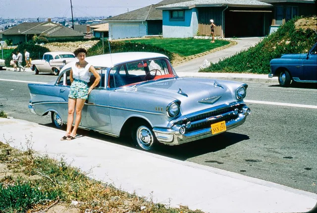 A woman in a white top and patterned shorts leans against a vintage silver 1957 Chevrolet parked on a suburban street, with houses and other cars visible in the background.