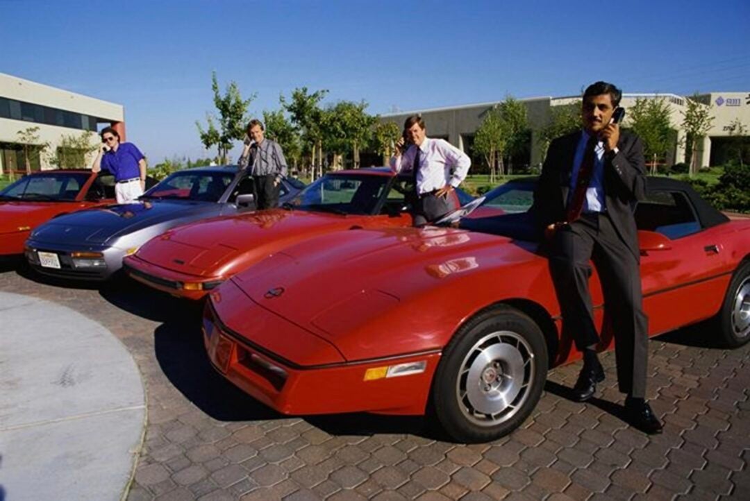 Four people in business attire pose confidently beside red sports cars parked on a brick driveway outside a modern office building; one man is talking on a mobile phone.
