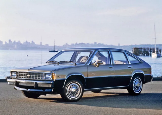 A vintage silver and black hatchback car is parked on a waterfront road, with calm water, sailboats, and a city skyline visible in the background.