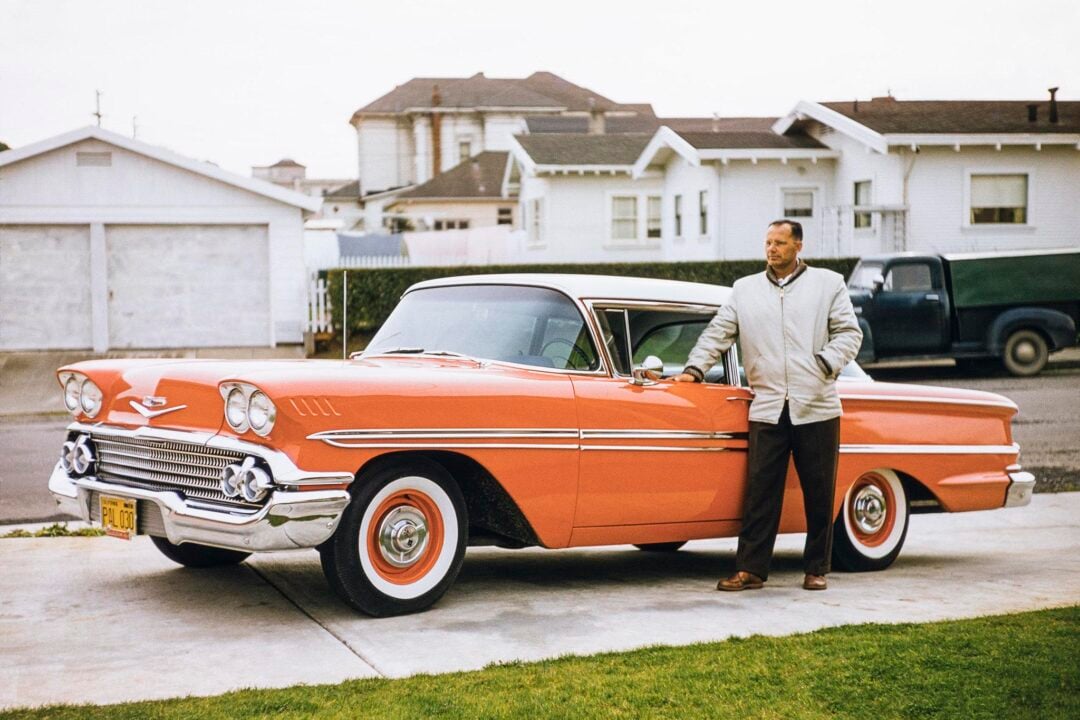 A man in a light jacket stands beside a classic orange 1950s Chevrolet car parked on a driveway in front of white houses and garages in a suburban neighborhood.
