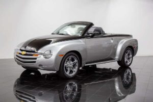 A silver and black Chevrolet SSR convertible is displayed in a showroom with a shiny black tile floor and a white background. The vehicle features chrome wheels and a retro-styled design.