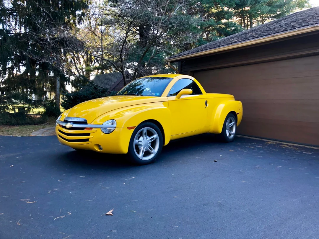 A bright yellow retro-style pickup truck is parked on a black asphalt driveway beside a brown garage, with trees and greenery in the background.