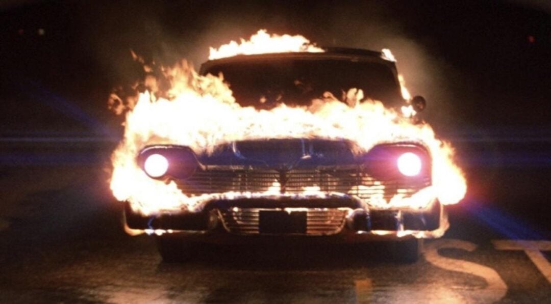 A vintage car is engulfed in bright flames, with its headlights shining through the fire as it drives on a dark, wet road at night.