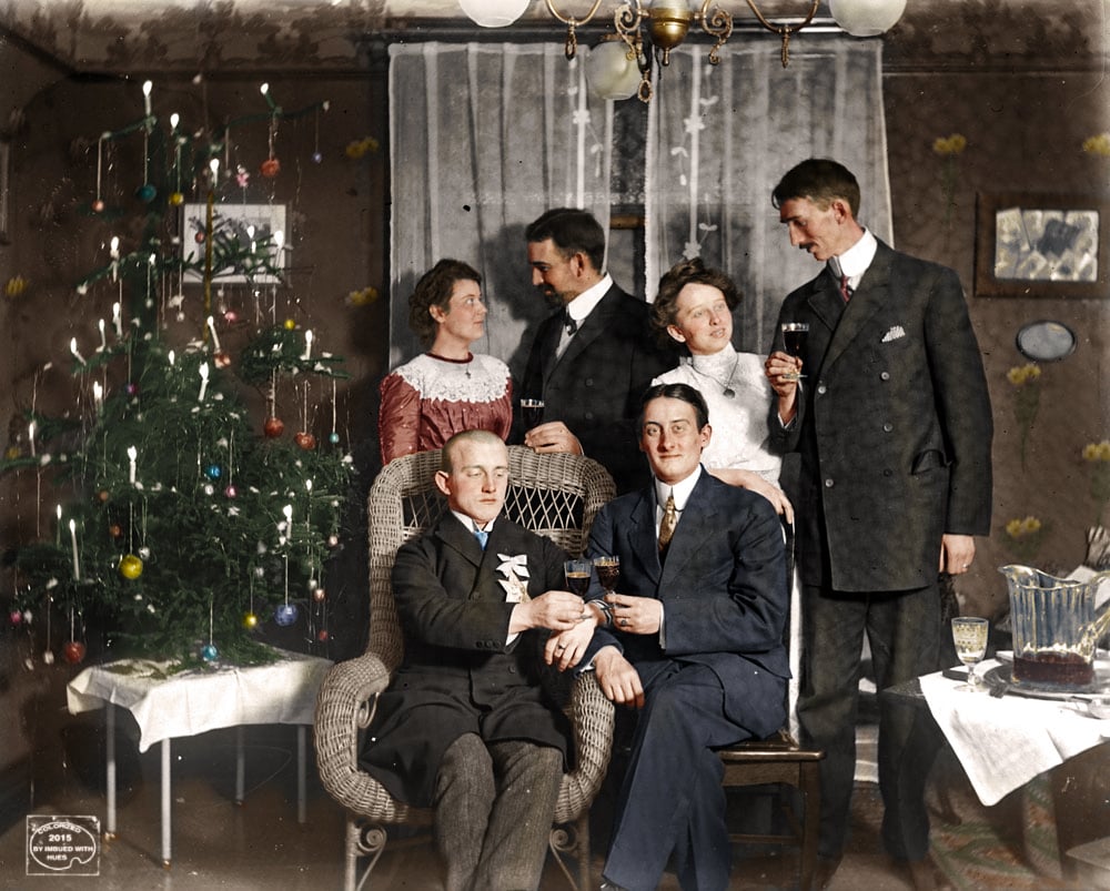 Six well-dressed adults gather in a decorated room by a Christmas tree, holding drinks and smiling. Two men sit front, four stand behind, with festive decorations, a chandelier, and wrapped gifts nearby.