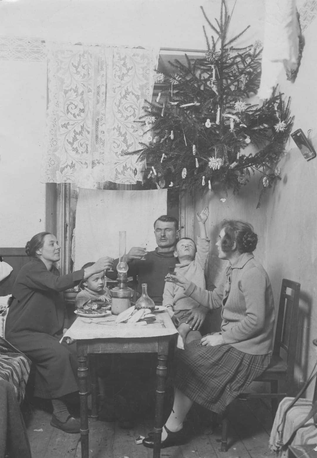A vintage black-and-white photo shows a family of four sitting around a small table, sharing a meal. An upside-down Christmas tree hangs from the ceiling, decorated with candles and ornaments.