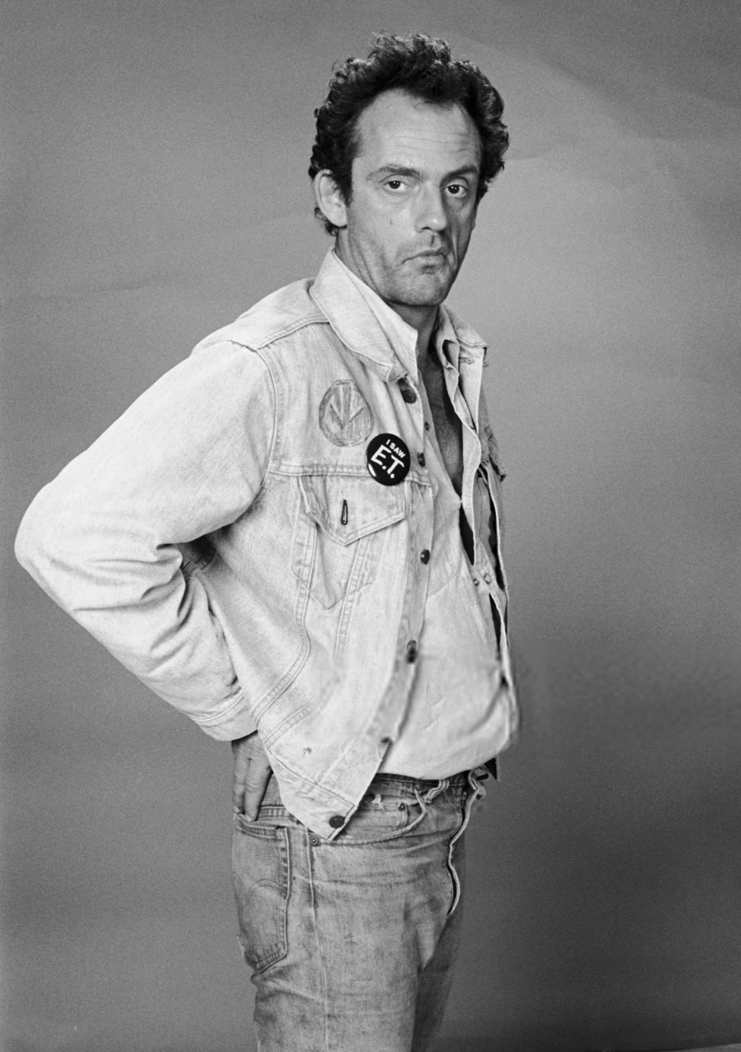 A man with short curly hair wears a light denim jacket and jeans, standing against a plain background. He looks seriously at the camera with one hand tucked behind his back. A round button is pinned to his jacket.