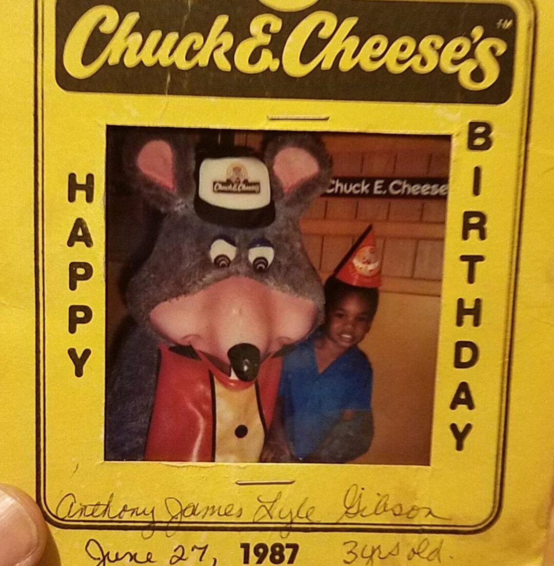 A hand holds a yellow Chuck E. Cheese’s birthday card featuring a photo of a child in a party hat standing next to the Chuck E. Cheese mascot. The card is signed and dated June 27, 1987, marking the child's 3rd birthday.