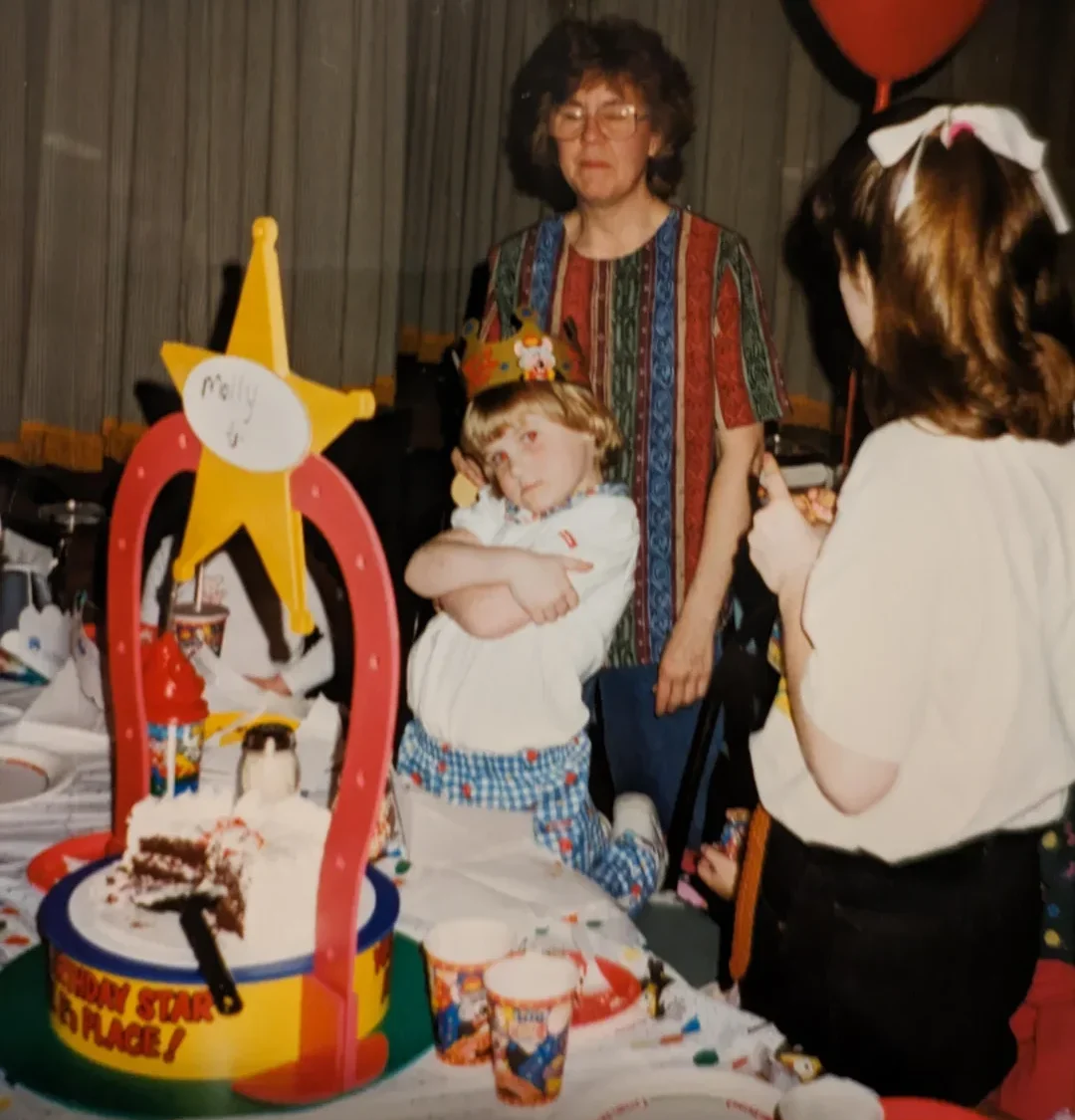 A young child wearing a birthday crown poses with crossed arms at a party table with cake and decorations, accompanied by an adult and another child holding a red balloon. The table is covered with party items and a partially eaten cake.